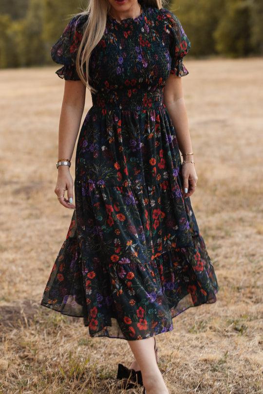 A woman wearing the Ivy City Co Cordelia Dress in Evergreen Vivid Meadow—a floral midi dress with puff sleeves, a smocked bodice, and a maternity-friendly fit—walks outdoors on dry grass with trees in the background. Her head is not visible in the image.