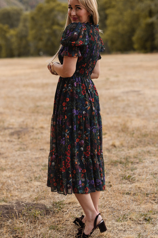A woman with long hair stands outdoors on dry grass, wearing the Cordelia Dress in Evergreen Vivid Meadow by Ivy City Co—a dark floral-patterned dress featuring a smocked bodice that is both elegant and maternity-friendly. She looks over her shoulder, smiling softly, with blurred trees in the background.