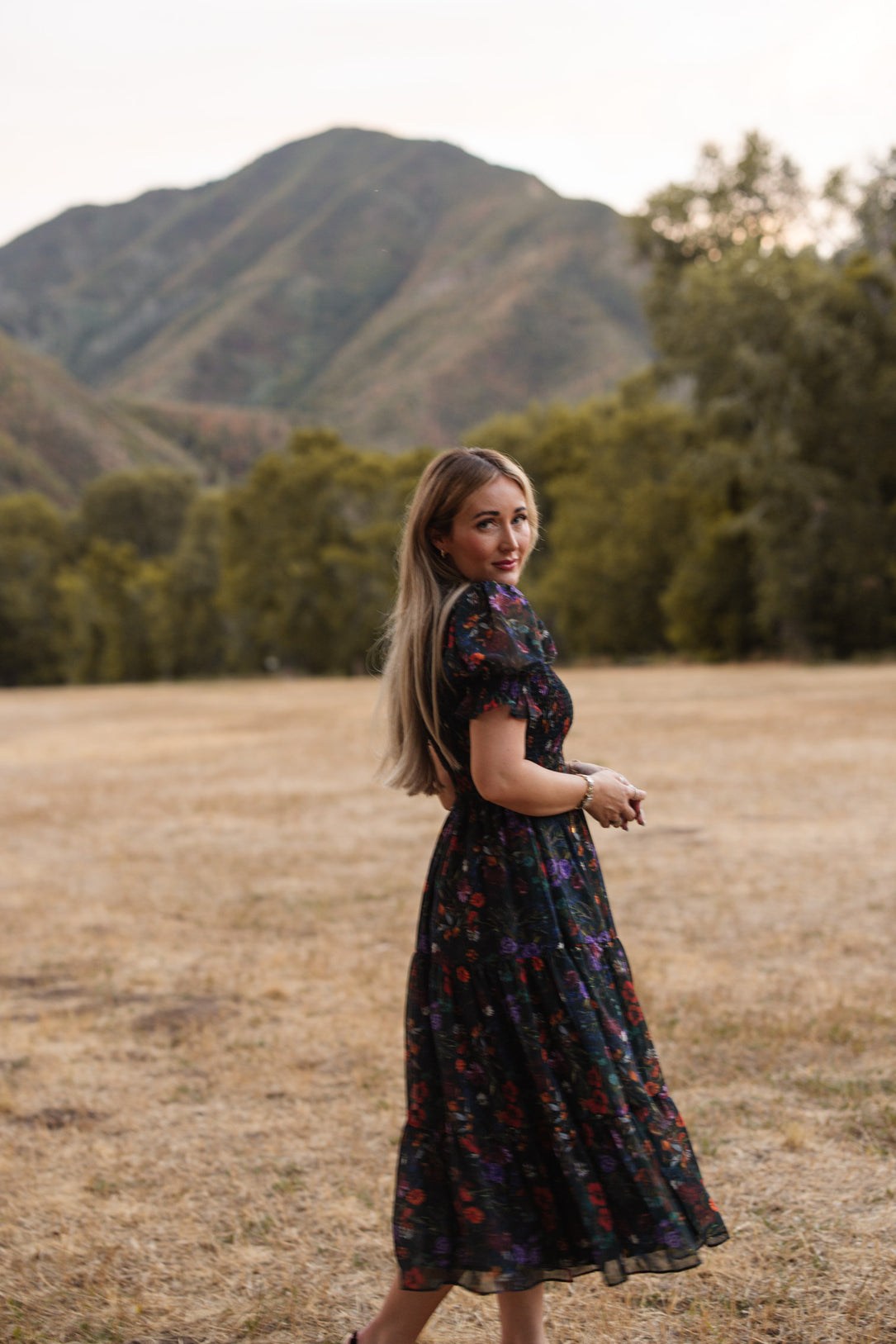 A woman wearing the Cordelia Dress in Evergreen Vivid Meadow by Ivy City Co, featuring a long, dark floral design and smocked bodice, stands in a dry grassy field looking back over her shoulder. Maternity friendly and flowing gracefully, the dress is set against a backdrop of green trees and a distant mountain.