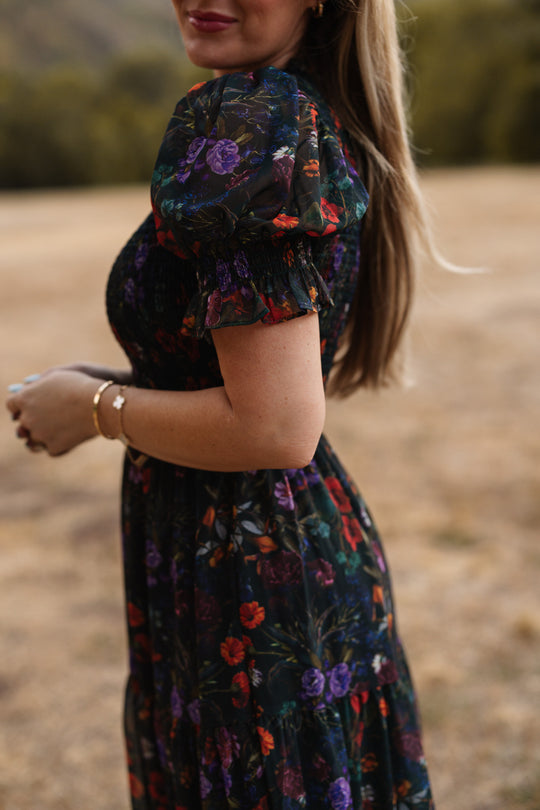 A woman stands outdoors on a grassy field wearing the Ivy City Co Cordelia Dress in Evergreen Vivid Meadow, which features a floral print, puffed sleeves, and a maternity-friendly smocked bodice. Her long hair is down, and she accessorizes with a bracelet. The softly blurred background showcases trees and rolling hills.