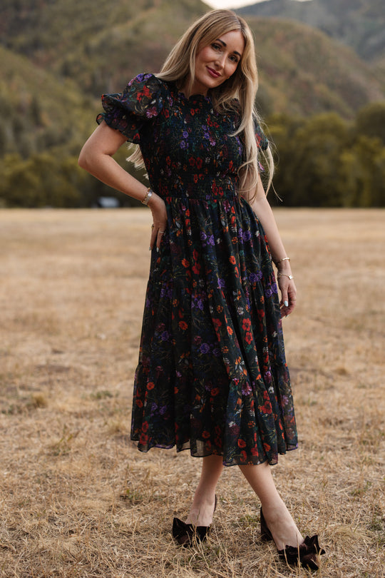 A woman stands outdoors on dry grass, wearing the Ivy City Co Cordelia Dress in Evergreen Vivid Meadow—a dark floral design featuring a smocked bodice and puff sleeves, paired with black bow heels. Green trees and mountains blur softly in the background as she smiles, her long blonde hair loose. This look is both elegant and maternity-friendly.
