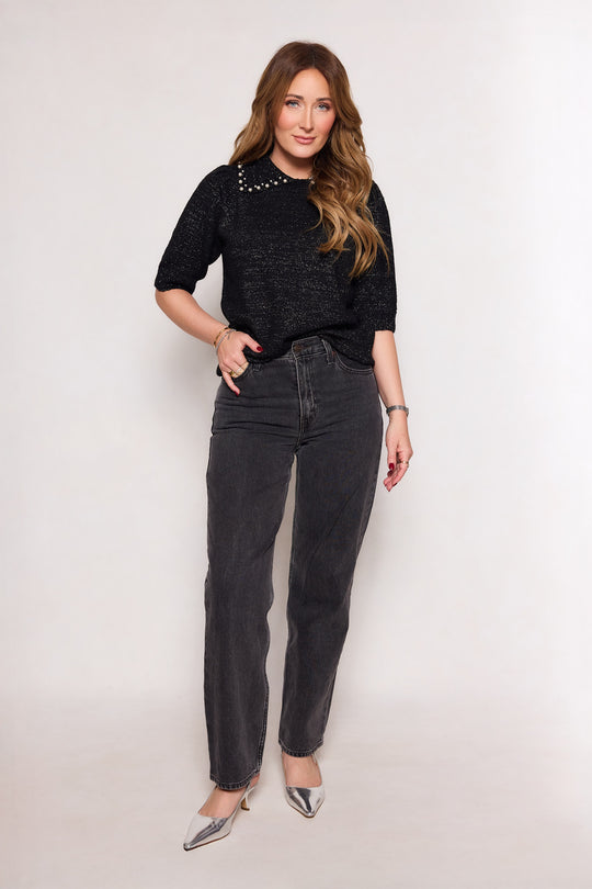 A woman with wavy brown hair stands against a plain background, wearing the Ivy City Co Corinth Sweater in Black with pearl details, high-waisted gray jeans, silver pointed-toe heels, and jewelry on her wrist and fingers.