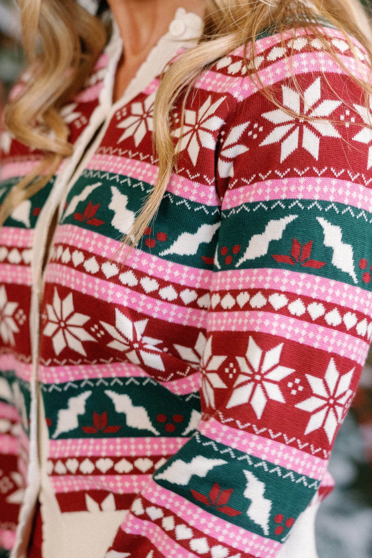 A close-up shows a person wearing the Ivy City Co Cornelia Cardigan in Festive Fair Isle, featuring red, pink, white, and green Fair Isle patterns with snowflakes, holly, and geometric designs. The person’s face is not visible. This cardigan is both maternity- and nursing-friendly for extra comfort during the holiday season.