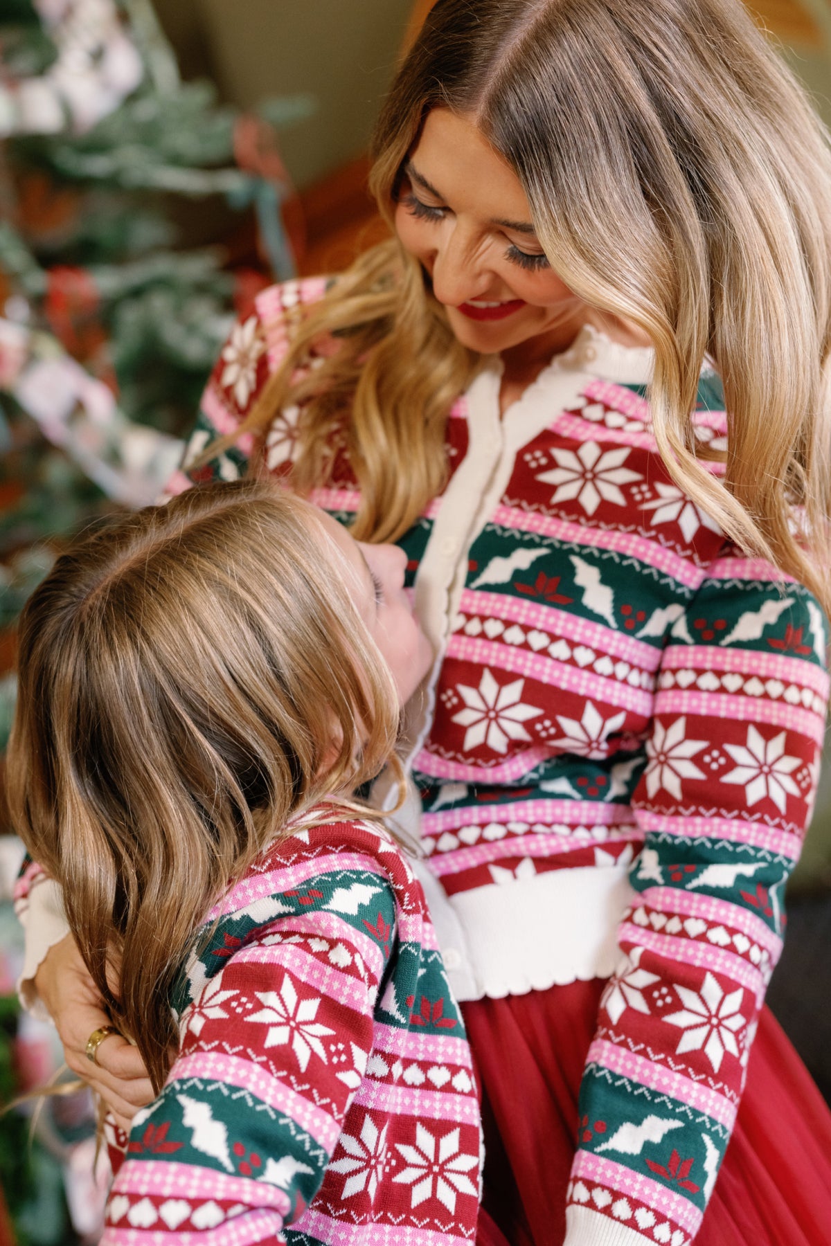 A woman and a young girl, both wearing matching Ivy City Co Cornelia Cardigan in Festive Fair Isle, smile and embrace in front of a decorated tree.