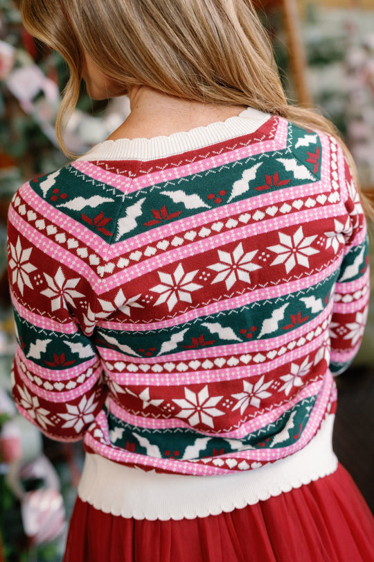 A person with long blonde hair, seen from behind, wears the Cornelia Cardigan in Festive Fair Isle by Ivy City Co—a festive red, white, green, and pink cotton sweater featuring snowflake and holly patterns. This Fair Isle cardigan includes a white collar and waistband, and is designed to be both maternity and nursing friendly.