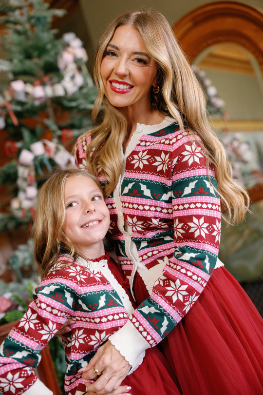 A woman and young girl, both dressed in matching Ivy City Co Cornelia Cardigan in Festive Fair Isle and red skirts, smile and hug in front of a decorated Christmas tree. These cozy cardigans are designed to be maternity and nursing friendly, making them perfect for family celebrations.
