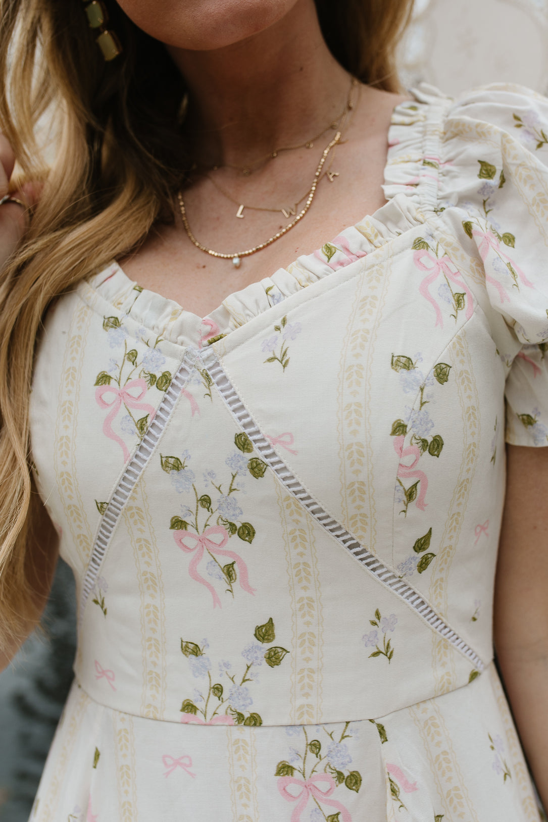 A woman wears the Ivy City Co Cottage Dress, a cream-colored piece with puff sleeves, pink ribbons, green leaf botanical print, and lace-trimmed sweetheart neckline. She pairs it with layered gold necklaces above her long blond hair.