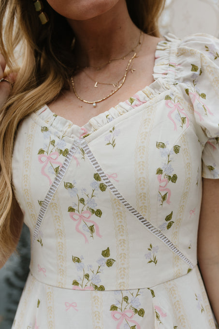A woman wears the Ivy City Co Cottage Dress, a cream-colored piece with puff sleeves, pink ribbons, green leaf botanical print, and lace-trimmed sweetheart neckline. She pairs it with layered gold necklaces above her long blond hair.