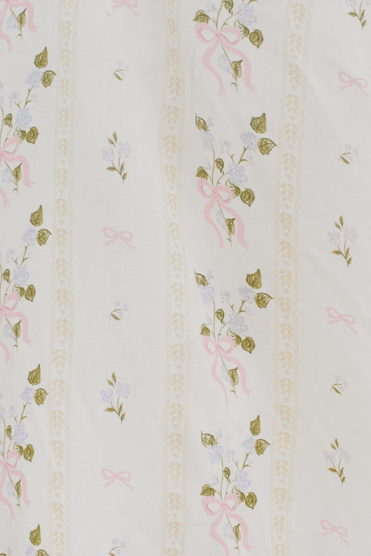 The Cottage Dress by Ivy City Co features white fabric adorned with small lavender flowers, green leaves, pink bows, and beige vine-like stripes—ideal for a charming cottage look.