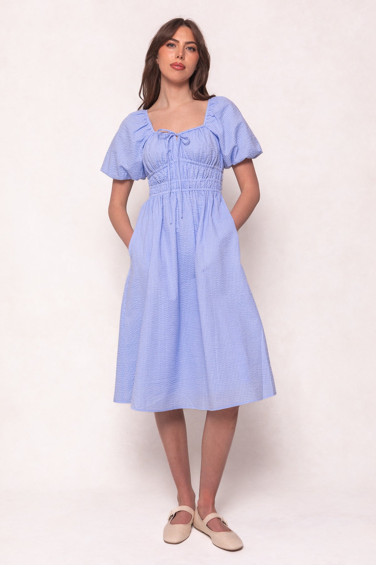 A woman models Ivy City Co’s Countryside Dress in Blue Seersucker—maternity friendly with short sleeves, smocked waist, tie detail, and pockets—against a plain background, finished with beige flats.