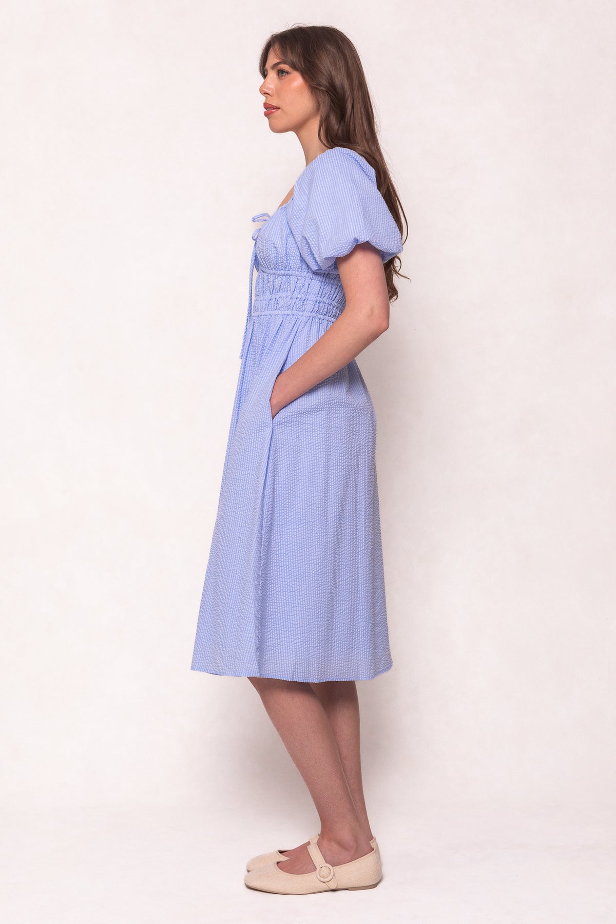 A woman stands in profile against a light background, wearing the Ivy City Co Countryside Dress in Blue Seersucker—short sleeves, fitted waist, and pockets make it a maternity-friendly choice. She has long brown hair and wears beige flats.