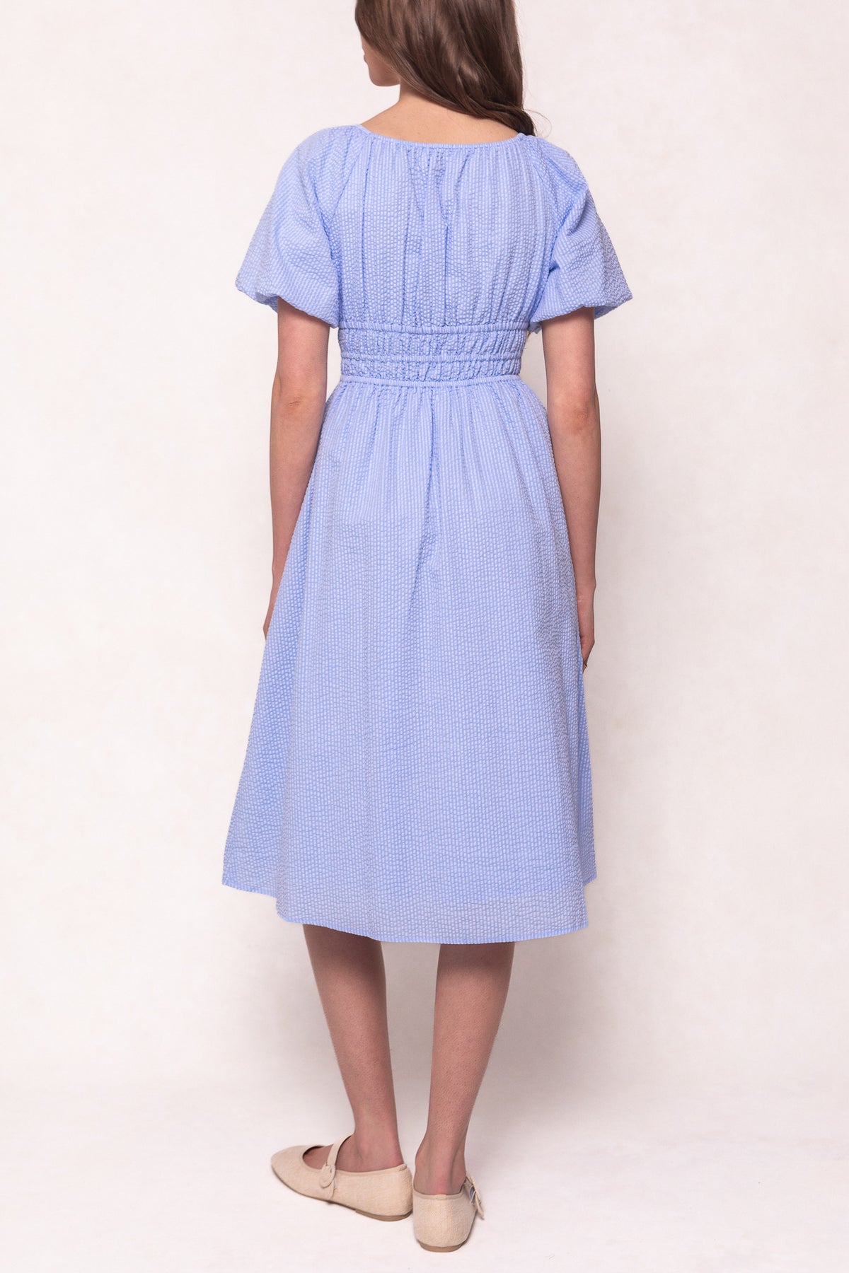 A woman with light skin and brown hair down wears the Ivy City Co Countryside Dress in Blue Seersucker, featuring a smocked waist, short puffed sleeves, and a nursing-friendly design. She pairs it with beige flats and faces away from the camera.