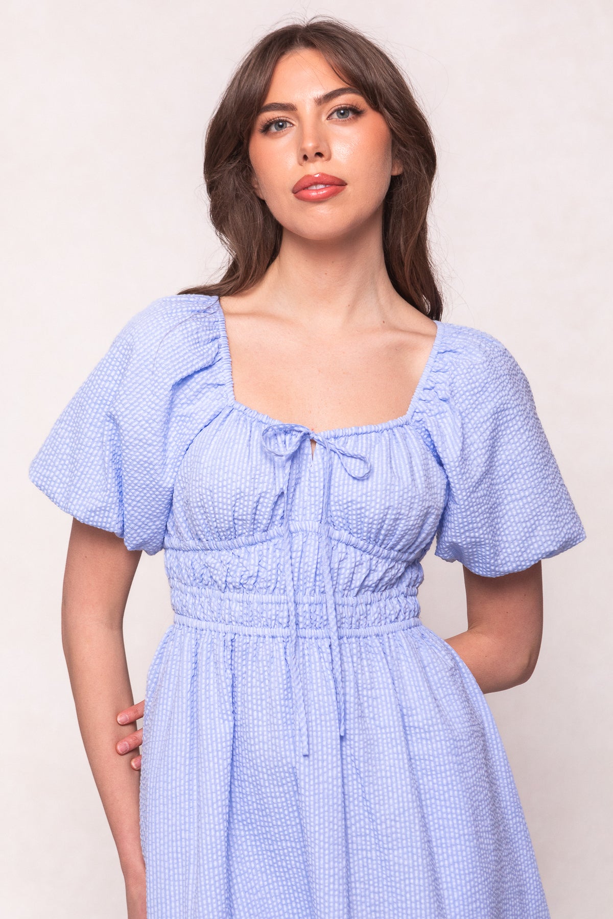 A woman with long brown hair wears the Ivy City Co Countryside Dress in Blue Seersucker—short-sleeved, with a square neckline and front tie—standing against a plain, light background.