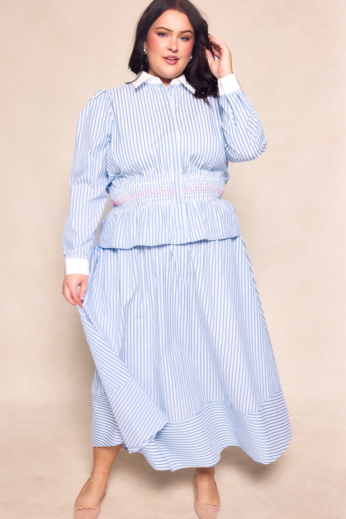 A woman poses confidently in Ivy City Co’s Bluebell Skirt in Blue & White Stripes paired with a matching long-sleeve blouse, featuring a white collar, cuffs, pink waistband detail, and nude heels—a maternity-friendly, effortlessly chic look.