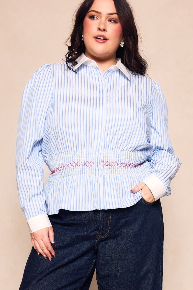 A woman models the Ivy City Co Bluebell Top in Blue & White Stripes, featuring white cuffs and collar, paired with dark jeans, posing with one hand in her pocket against a beige background.