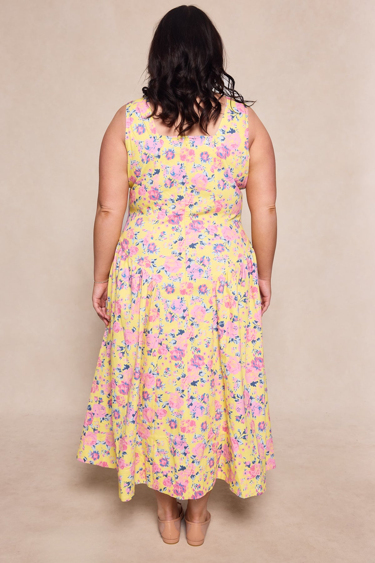 A woman with dark hair stands facing away, wearing the Ivy City Co Edith Dress in As You Dream Floral—a sleeveless yellow dress with pink and purple flowers—and beige shoes, set against a neutral background.