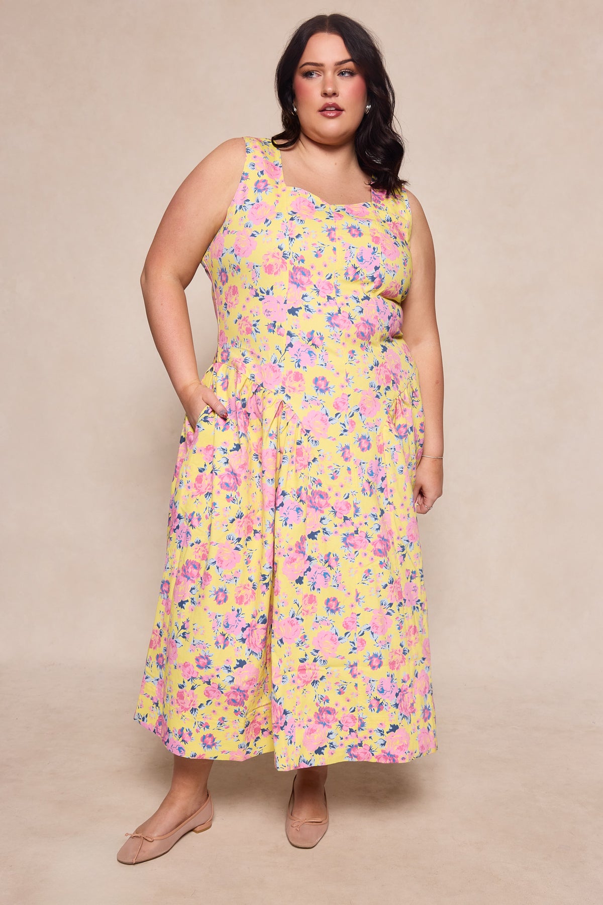 A woman with dark hair wears the Ivy City Co Edith Dress in As You Dream Floral, a sleeveless yellow dress with pink and purple flowers. She stands against a beige background in nude flats, posing with one hand in her pocket.
