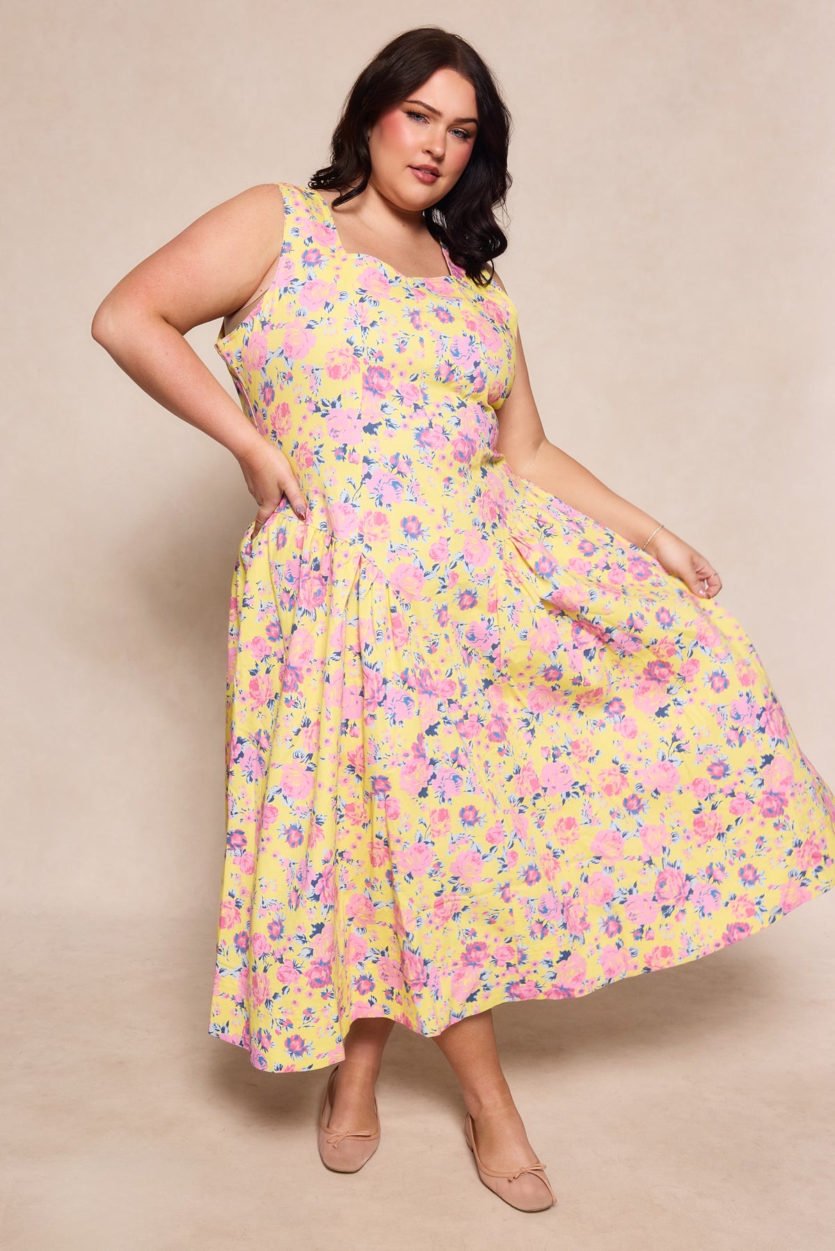 A woman models the Edith Dress in As You Dream Floral by Ivy City Co, a sleeveless yellow floral spring dress. She poses against a light beige background, holding out its flowy skirt and pairing it with nude flats.