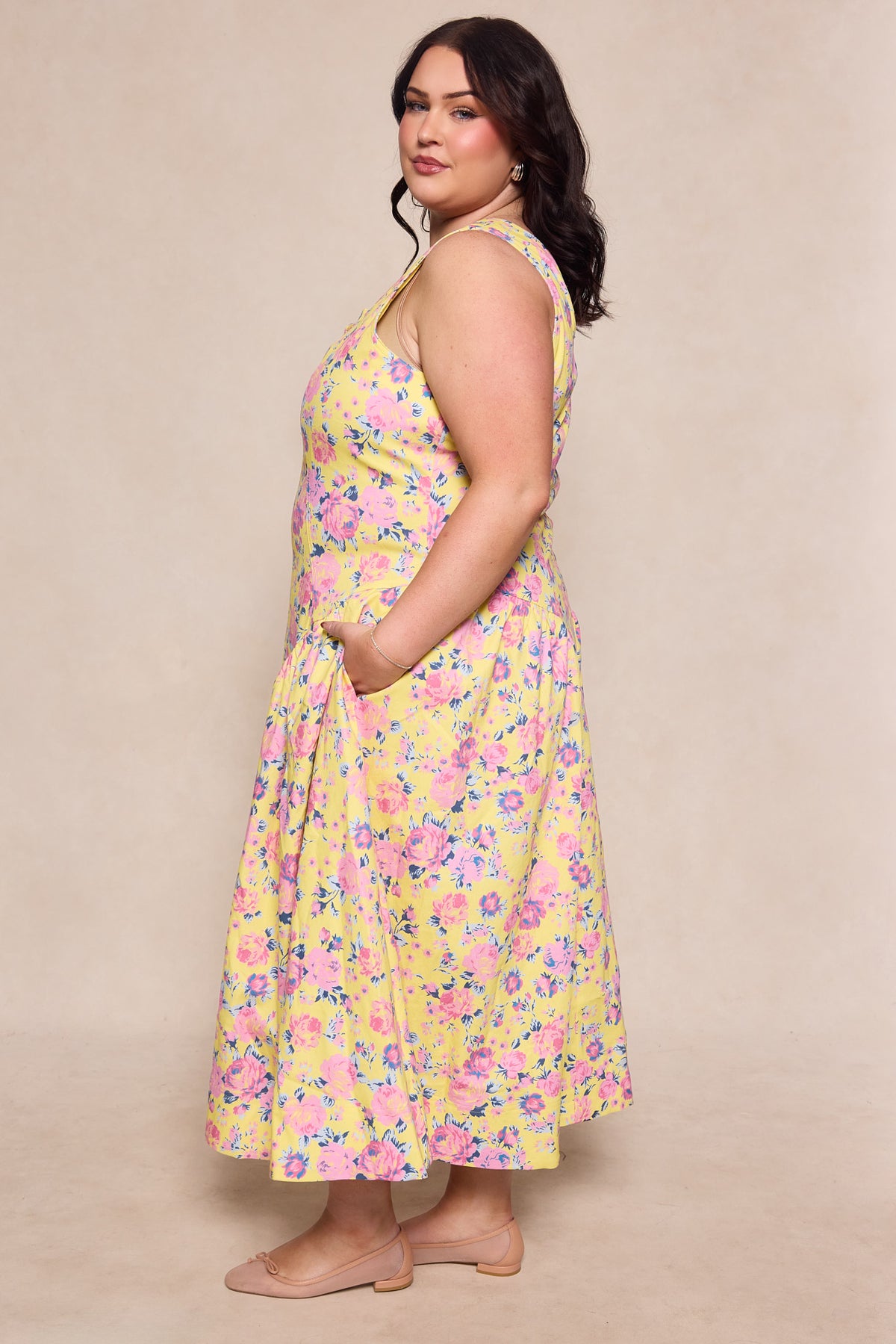 A woman wearing the Ivy City Co Edith Dress in As You Dream Floral, a sleeveless yellow spring dress with floral print and pockets, stands in profile against a neutral background with long dark hair and nude flats.
