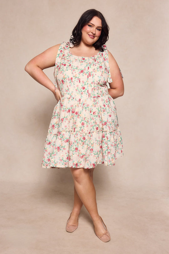 A woman wearing the Ivy City Co Helene Dress in Vibrant Chintz, a sleeveless, knee-length floral dress, and nude flats poses with one hand on her hip against a beige background.