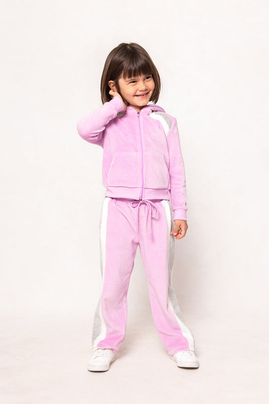 A young child with short brown hair wears the Ivy City Co Mini Kailey Zip Up Velour Sweatshirt in Pink and white sneakers, standing against a plain white background, smiling with one hand touching their head.
