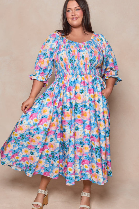 A woman in the Ivy City Co Teagan Dress in Delia Blue—a vibrant, floral midi dress featuring long puffed sleeves, a smocked cotton bodice, and a gathered waist—smiles and poses against a neutral background. She pairs the look with white sandals.