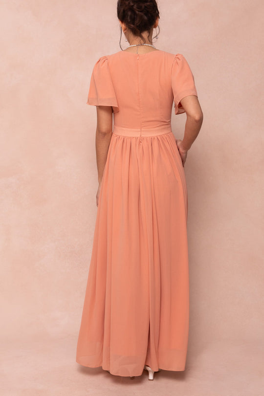 A woman stands facing away, wearing the Abbie Dress in Apricot Crush by Ivy City Co—a maxi dress featuring short sleeves, a fitted waist, and a flowing skirt—set against a light pink textured background.