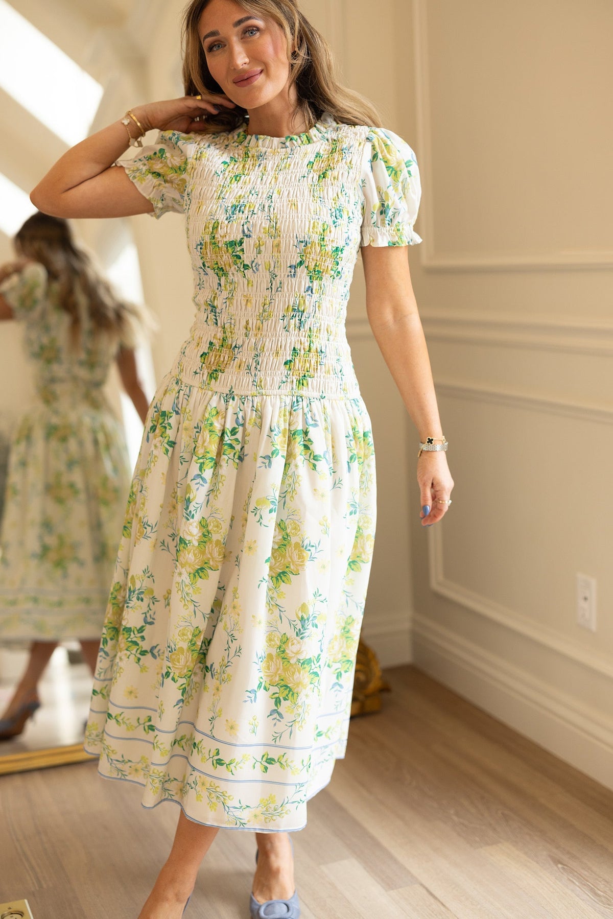 A woman smiles as she stands on a wooden floor in a well-lit room, wearing the Ivy City Co Jennifer Dress in Garden Tea Party Floral. This white floral print dress features short puff sleeves, is crafted from lightweight fabric, and is designed to be maternity friendly. The mirror behind her reveals both the front and back views of the dress.