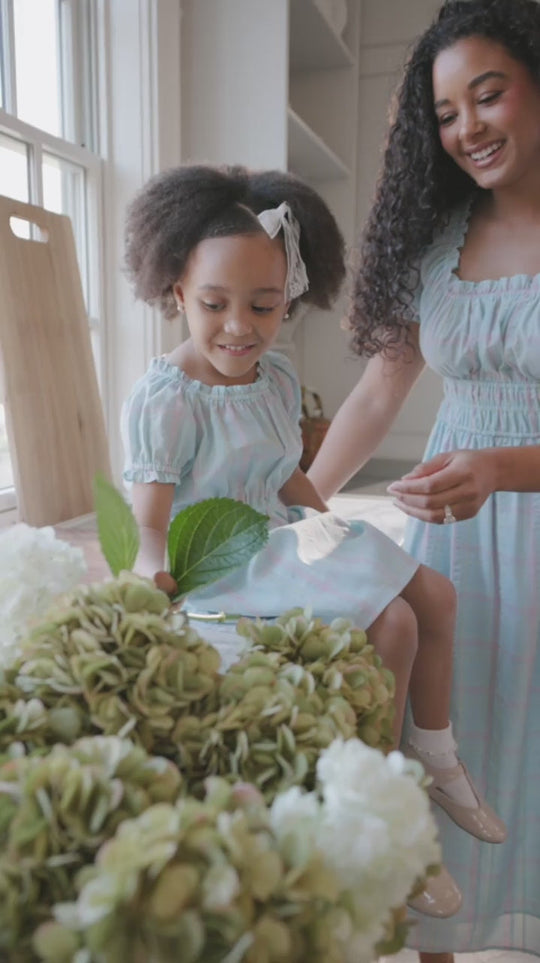 A woman and a young girl smile together indoors near a bouquet of green and white flowers, both wearing the Ivy City Co Thalia Dress in Pastel Plaid.