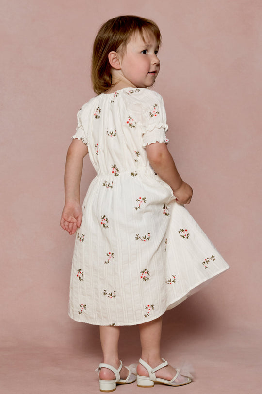 A young child stands on a pink background, looking over her shoulder, wearing the Ivy City Co Mini Jessie Dress in Ditsy Embroidery—a cream-colored dress adorned with small floral patterns. She completes the look with white sandals and has light brown hair.