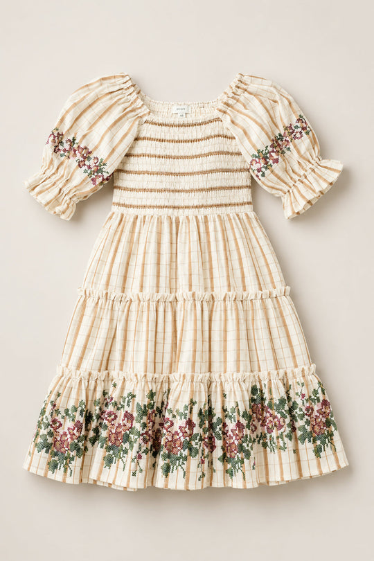 The Mini Madeline Dress in Embroidered Cider Grid by Ivy City Co features a cream and beige plaid pattern with puff sleeves, a smocked bodice, and delicate floral embroidery on the sleeves and hem. The skirt is finished with ruffled tiers adorned with green, pink, and burgundy flowers.