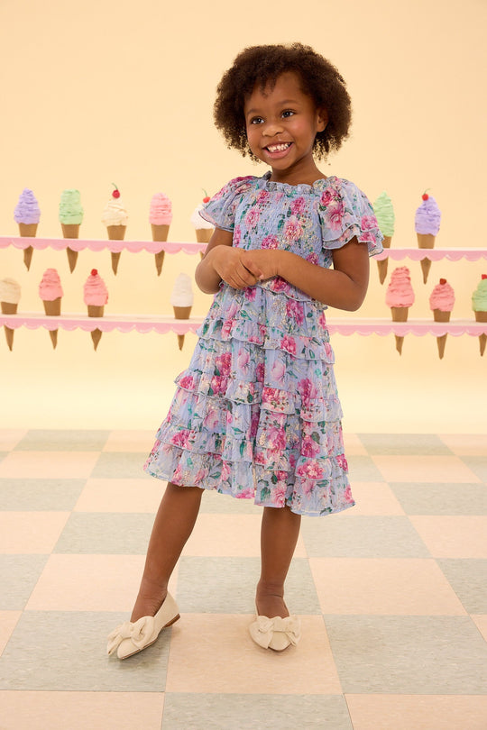 A smiling young girl wears the Ivy City Co Mini Winnie Dress in Peony Blossoms, paired with bow shoes, as she stands on a checkered floor with colorful ice cream cone decorations adorning the wall behind her.