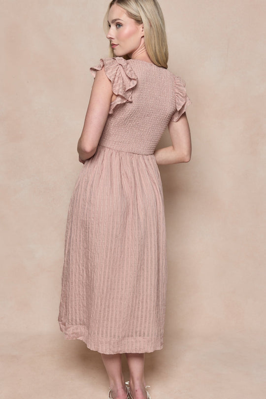 A woman with long blonde hair wears the Hattie Dress in Pink Stripes by Ivy City Co—a light pink, knee-length dress featuring soft pink stripes, ruffled sleeves, and a smocked bodice for a fresh, feminine look. Maternity friendly, she poses against a beige backdrop with her back partially turned to the camera.
