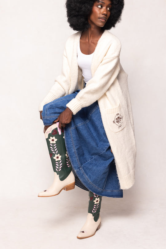 Woman wearing a long cream cardigan, blue denim skirt, and patterned leggings on a white background