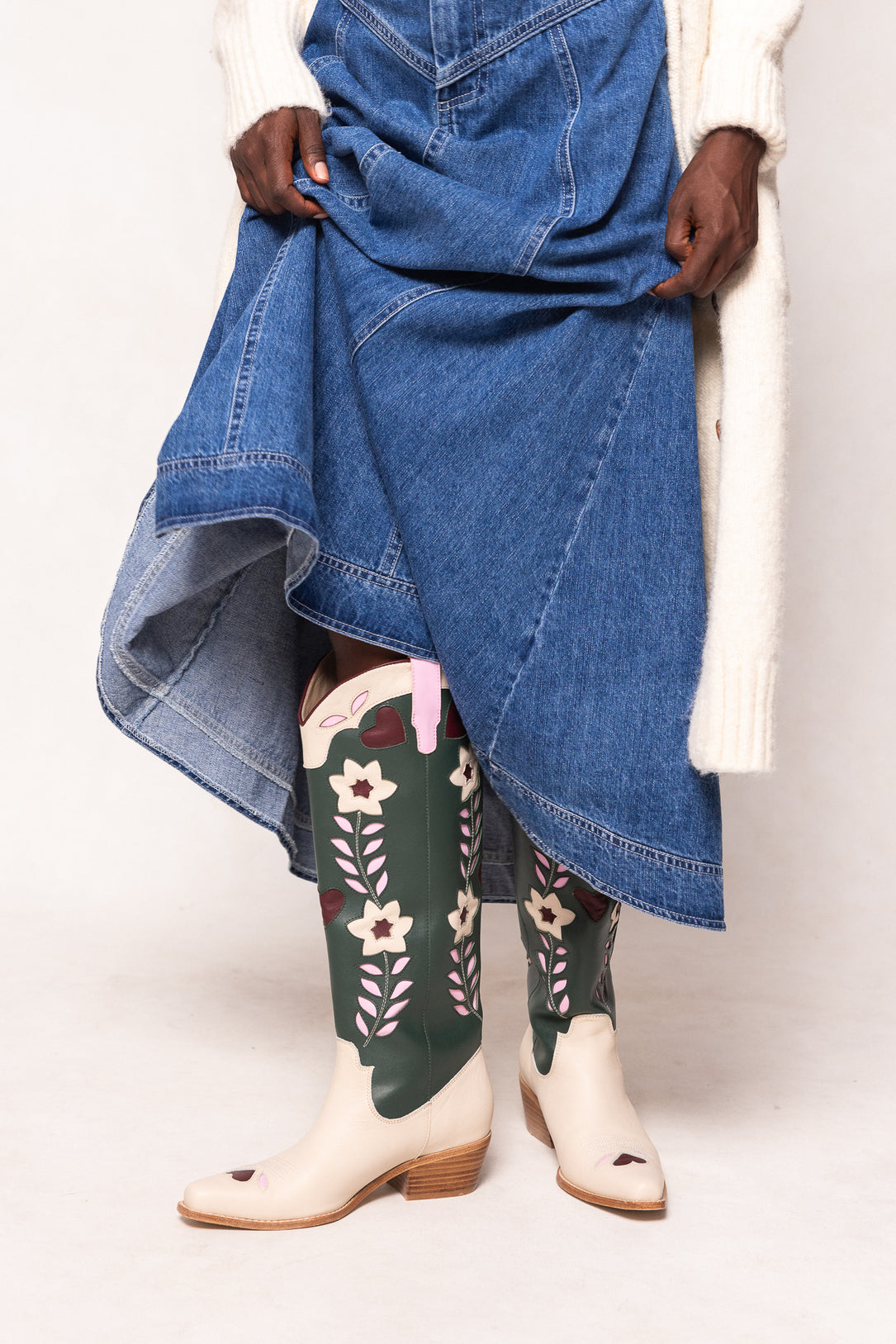Person wearing a blue denim skirt, green floral boots, and white shoes on a white background