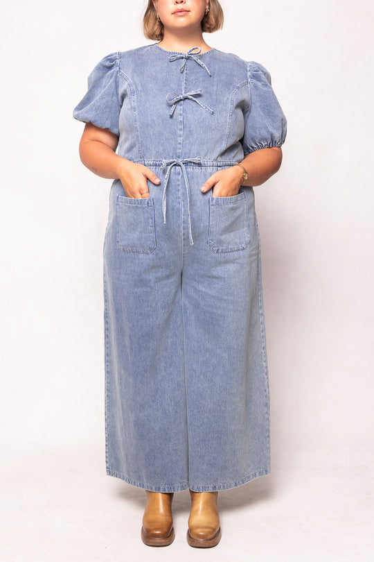 A person wears the Dallas Jumpsuit in Denim by Ivy City Co, featuring puff sleeves, front ties, large pockets, and wide legs. Their hands are in the pockets and they finish the look with tan heeled shoes.
