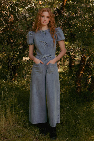 A woman with long red hair stands outdoors in a sunlit, wooded area wearing the Ivy City Co Dallas Jumpsuit in Denim, featuring short puffed sleeves, front pockets, and bows, paired with black boots.