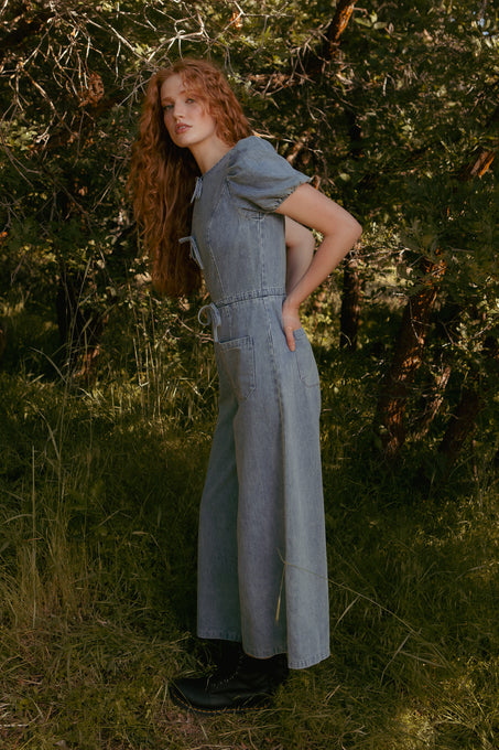 A woman with long red hair stands sideways in a sunlit, wooded area, wearing the Ivy City Co Dallas Jumpsuit in Denim and black boots. She looks toward the camera with a relaxed expression among trees and green grass.