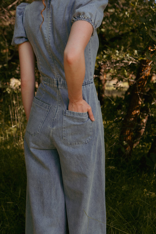 A person wears the Dallas Jumpsuit in Denim by Ivy City Co outdoors, with one hand in a back pocket. Trees and sunlight can be seen in the background.