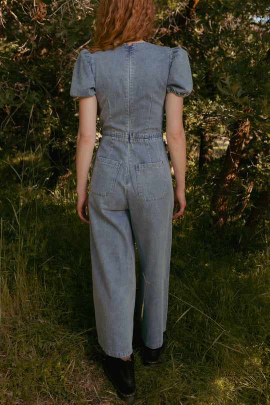 A person with wavy, shoulder-length hair stands on grass facing away, wearing Ivy City Co’s Dallas Jumpsuit in Denim—light blue with wide legs and short puff sleeves—paired with black boots, surrounded by tall green trees and foliage.