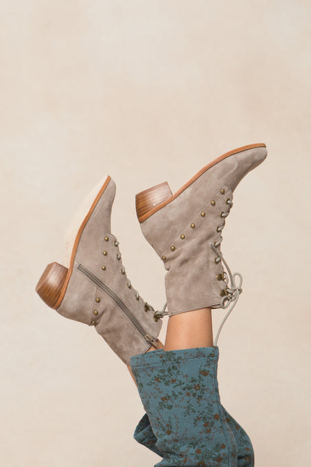 A person in green floral pants lifts their legs, highlighting the Ivy City Co Mini Darcy Lace Up Studded Boot—tan suede with wooden heels and metal studs—set against a neutral background.