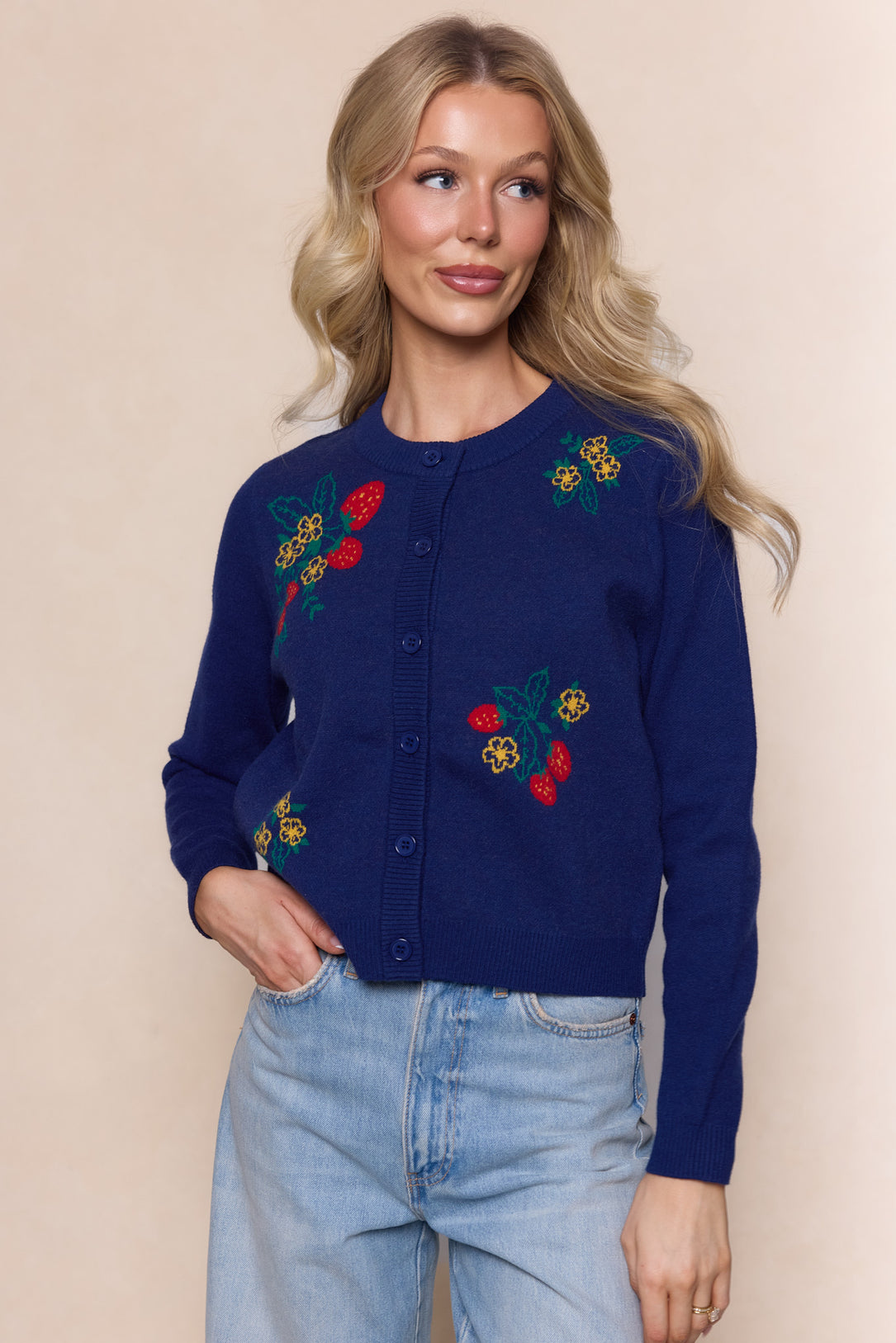 A woman with long blonde hair wears the Ivy City Co Delphi Sweater in Strawberry Print, featuring colorful floral and fruit embroidery, paired with light blue jeans against a plain beige background.