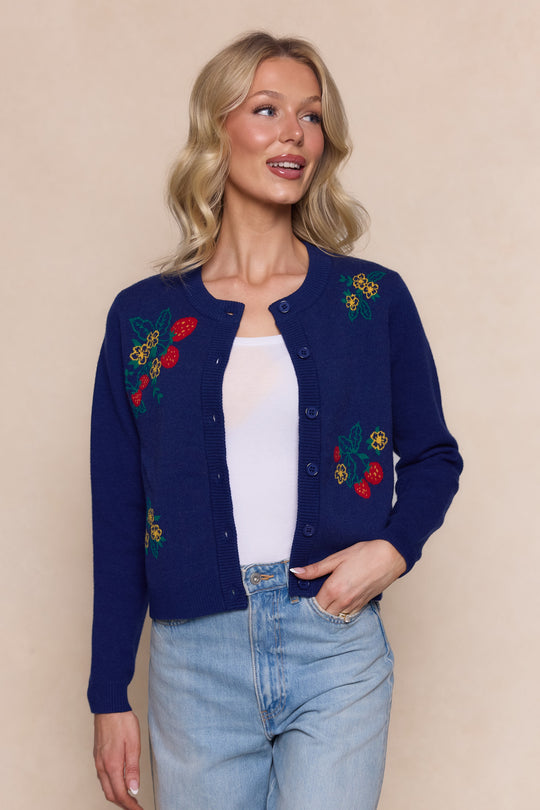 A woman wearing the Delphi Sweater in Strawberry Print by Ivy City Co—a navy knit cardigan with colorful embroidered berries—over a white top and light blue jeans, stands against a beige background, smiling and looking to the side.