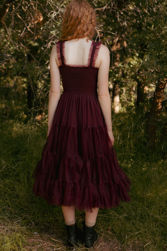 Person wearing a burgundy dress with ruffled layers standing in a forest.