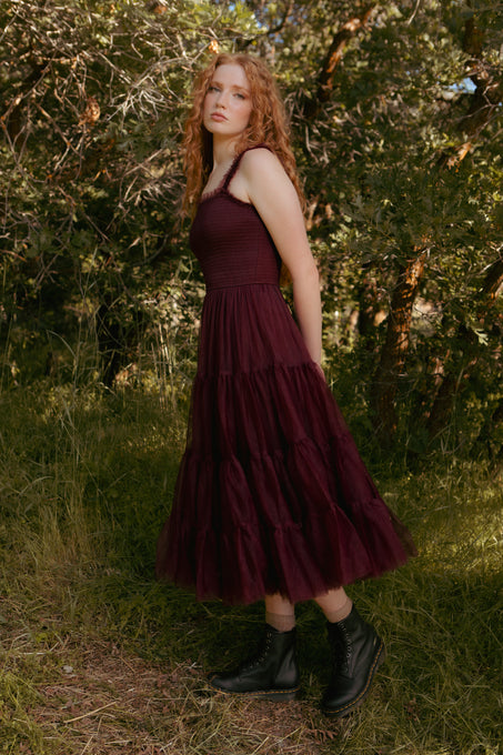 Woman in a burgundy dress standing in a natural setting with greenery.