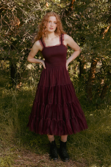 Woman wearing a burgundy dress with ruffled layers standing in a natural setting.