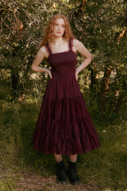 Woman wearing a burgundy dress with ruffled layers standing in a natural setting.