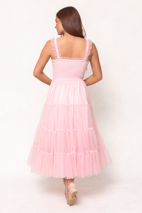 A woman with long brown hair stands with her back to the camera in the Ivy City Co Destiny Dress in Pink, featuring soft, tiered tulle layers. She wears nude platform heels against a light background, highlighting this feminine maternity style.