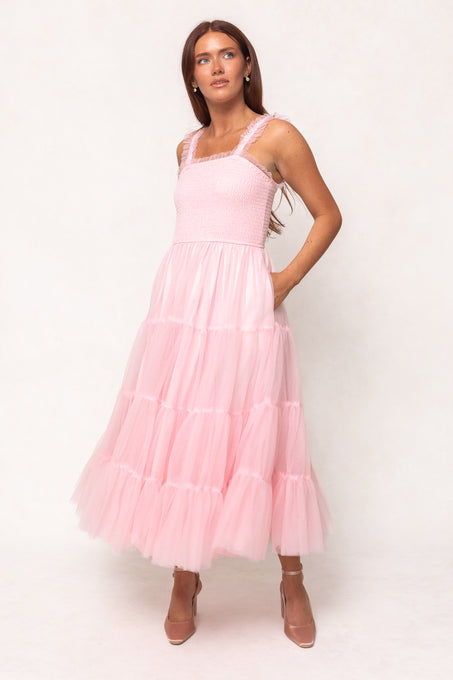 A woman in Ivy City Co’s Destiny Dress in Pink—a sleeveless, tiered tulle maternity dress with a smocked bodice and ruffled straps—stands against a light background, paired with pink heels. Perfect for special occasions.