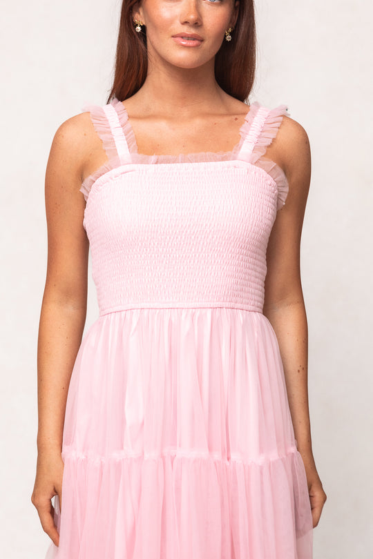 A woman wears the Ivy City Co Destiny Dress in Pink, featuring ruffled straps, a smocked bodice, and a flowy, tiered skirt. Set against a plain background, this maternity and nursing-friendly dress stands out for its charm and elegance.